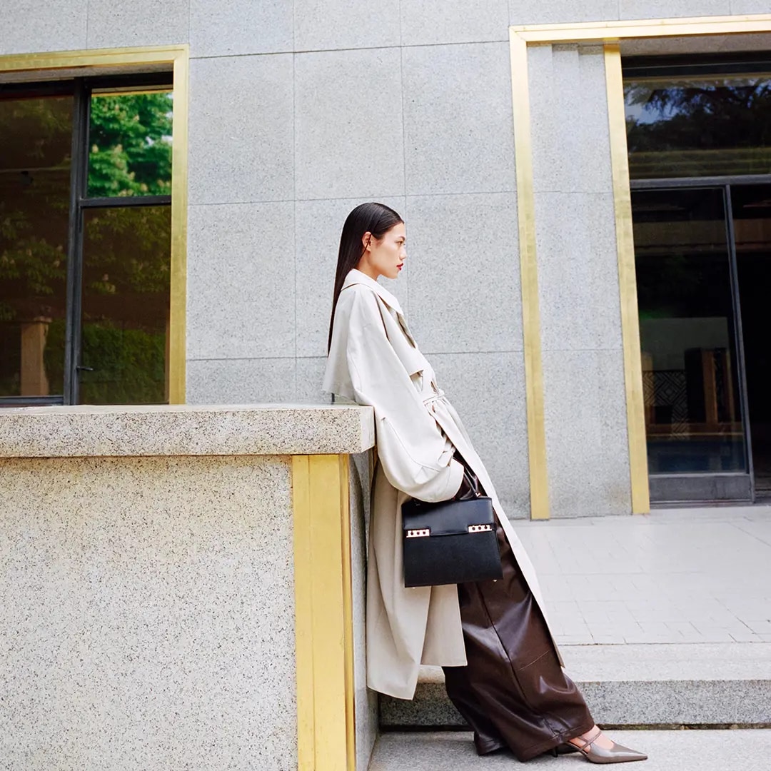 Model wearing a long white trench coat and leather brown pant with a Delvaux black hand bag
