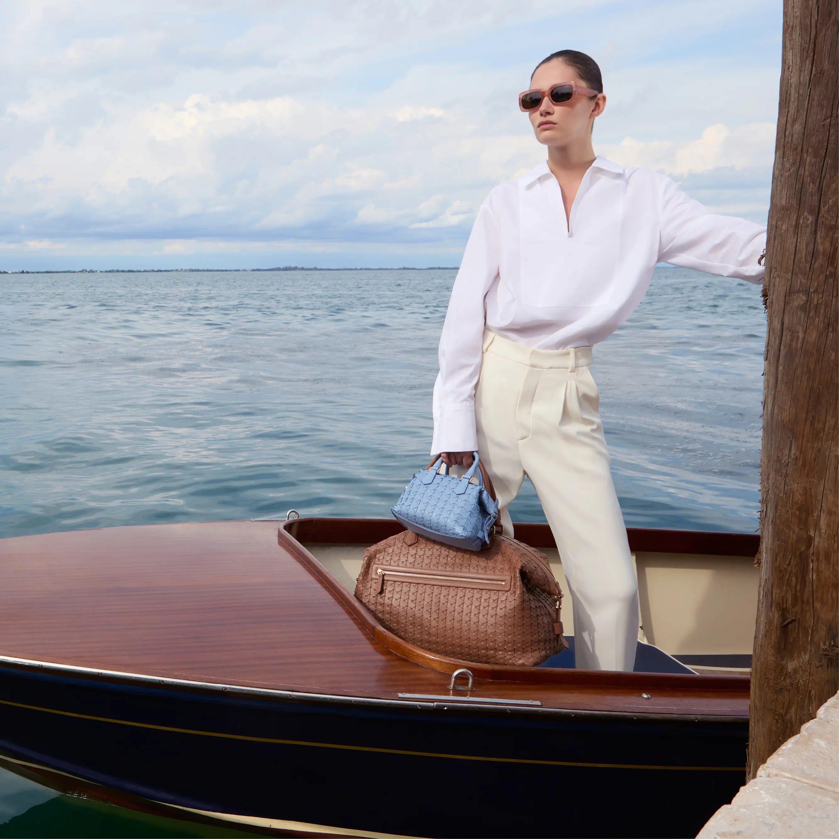 Model pausing in a speed boat holding blue small mosaic bad and large light brown mosaic bag