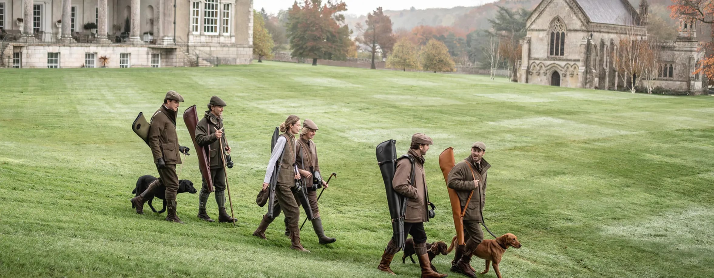 A group of people dressed in traditional hunting attire, accompanied by dogs, walking across a green lawn towards a grand estate and a smaller, church-like building.